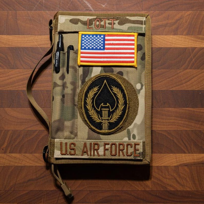 Pu&ograve; includere: Copertina per notebook con motivo mimetico con una toppa della bandiera americana e una toppa dell'US Air Force. La copertina ha un portapenne e una tracolla. Il nome "LOTT" &egrave; ricamato in alto.