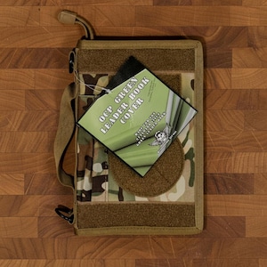 Pu&ograve; includere: Una copertina per libri con motivo mimetico con tracolla e gancio. La copertina ha una toppa a strappo e un'etichetta con la scritta "OCP GREEN LEADER BOOK COVER". La copertina &egrave; beige, verde e marrone.