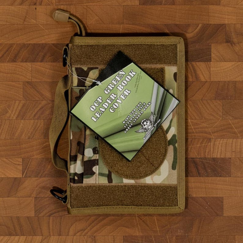 Pu&ograve; includere: Una copertina per libri con motivo mimetico con tracolla e gancio. La copertina ha una toppa a strappo e un'etichetta con la scritta "OCP GREEN LEADER BOOK COVER". La copertina &egrave; beige, verde e marrone.