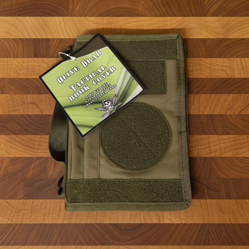 Pu&ograve; includere: Copertina per libri tattica verde oliva con tracolla. La copertina ha un'ampia toppa circolare e diverse toppe rettangolari. Il tag allegato recita "Olive Drab Tactical Book Cover" con testo aggiuntivo e un logo con teschio e ali.
