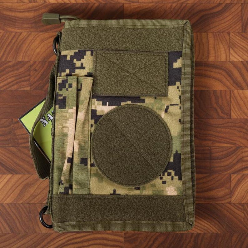 Pu&ograve; includere: Copertina per taccuino tattico mimetico verde e beige con tracolla e pi&ugrave; tasche. La copertina ha una grande toppa circolare e una toppa rettangolare per attaccare elementi aggiuntivi. Un taccuino con le lettere "NA" &egrave; visibile all'interno.