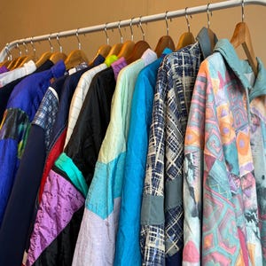 Może przedstawiać: Stojak z vintage'owymi kurtkami w różnych kolorach i wzorach, w tym niebieskim, fioletowym, zielonym i różowym. Kurtki są wykonane z nylonu i mają sportowy, retro wygląd.