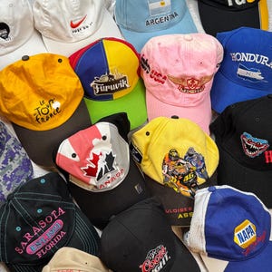 Peut inclure: Une collection de casquettes de baseball de différentes couleurs, dont le blanc, le bleu, le jaune et le noir. Plusieurs casquettes présentent des logos, du texte et des motifs, tels que "JUST DO IT", "ARGENTINA" et "SARASOTA EST. 1921 FLORIDA".