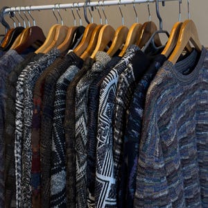 Puede incluir: Una colección de suéteres con estampados coloridos colgados en perchas de madera. Los suéteres son una mezcla de azul, marrón, blanco y negro.