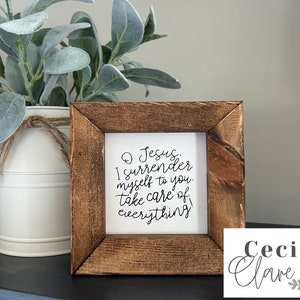 May include: A framed canvas sign with the text "O Jesus, I surrender myself to you. Take care of everything." The sign is in a rustic brown frame and is sitting on a black surface. The sign is from Cecilia Clare & Co.