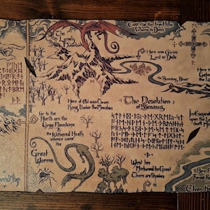 May include: A vintage-style map featuring illustrations of mountains, a dragon, and a river. The map includes text in English and a script, with the title "The Desolation of Smaug". The map is printed on a tan-colored material.
