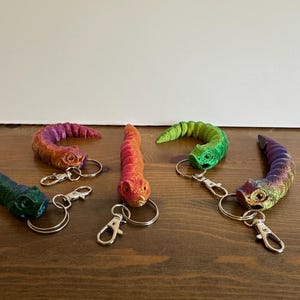 May include: Six colourful worm-shaped keyrings are displayed. Each keyring has a unique gradient of colours, including blue, green, orange, and purple. The keyrings are made of a shiny material and have silver-coloured key rings and clasps.