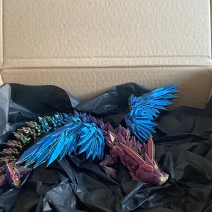 May include: A iridescent blue and purple origami dragon with wings spread wide, resting in a black tissue-lined cardboard box.