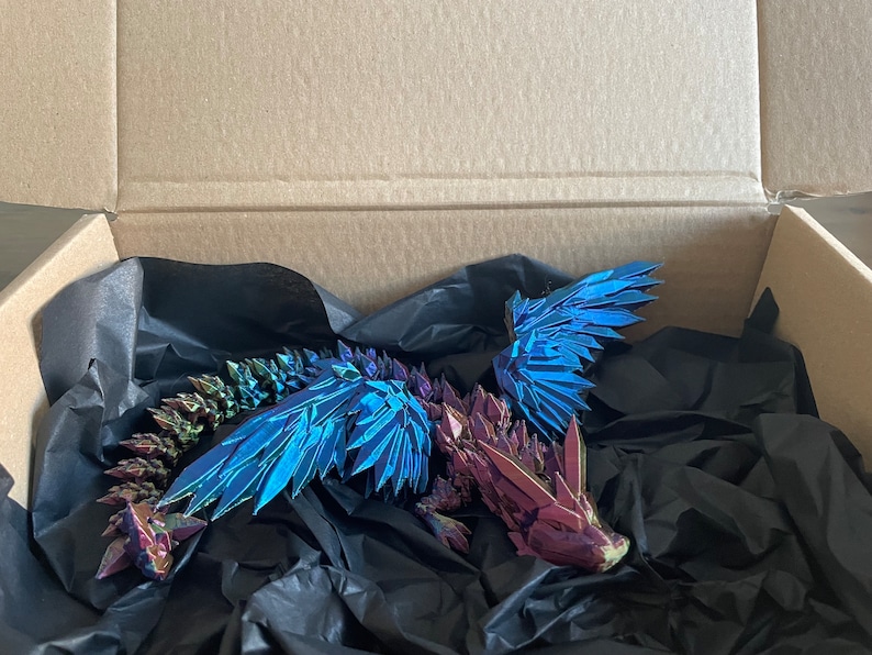 May include: A iridescent blue and purple origami dragon with wings spread wide, resting in a black tissue-lined cardboard box.