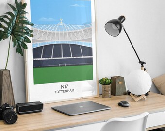 May include: A framed art print featuring a stylized illustration of a stadium with a green pitch, dark blue seating, and a light blue sky. The text N17 TOTTENHAM is printed at the bottom of the white border. The print is displayed in a light wood frame.