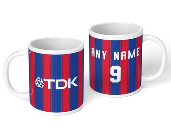 May include: Two white ceramic mugs featuring a vertical red and blue striped pattern. One mug displays the white TDK logo with a geometric icon. The other mug features the white text ANY NAME above the large white number 9 in a block font.