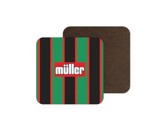 May include: A square drink coaster featuring a vertical striped pattern in black, green, and red. In the center, a red rectangular box contains the white text müller. A second, plain dark brown square coaster is partially visible behind the first.