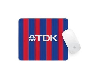 May include: A rectangular mouse mat featuring a vertical striped pattern in alternating shades of blue and red. The white TDK logo is printed in the centre of the mouse mat. A smooth white computer mouse is positioned on the right side of the mat.