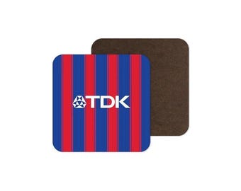 May include: A square coaster featuring a pattern of vertical blue and red stripes with the white TDK logo centred on the front. The coaster has a dark brown, textured underside visible behind the main striped surface.