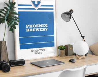 May include: A framed rectangular poster featuring a blue and white graphic design resembling a sports jersey. The text PHOENIX BREWERY is centred in white, with BRIGHTON 1985/86 printed below. The design includes horizontal white stripes and a shield.