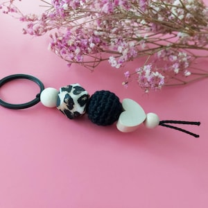 Può includere: Un portachiavi nero con un anello nero, su cui sono infilate una perla di legno con stampa leopardata bianco e nero, una perla di uncinetto nero, una perla di legno a forma di cuore bianco e una perla di legno bianca. Le perle sono attaccate a un cordino nero.