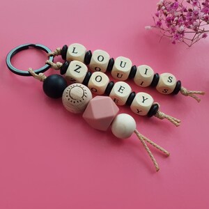 Può includere: Portachiavi personalizzati con nomi. I portachiavi presentano perline di legno con le lettere che compongono i nomi "LOUIS" e "ZOEY", insieme a perline decorative nere, rosa e in legno naturale. Sono attaccati a un anello di metallo nero.
