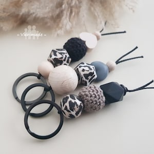 Può includere: Un portachiavi nero con una varietà di perline di legno e plastica, tra cui un cuore, una stampa leopardata e perline all'uncinetto. Il portachiavi ha anche un anello di metallo nero.