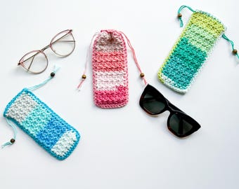 May include: An assortment of handmade crochet eyeglass cases in various colors, including pink, blue, and green. The cases are shown with a pair of eyeglasses and sunglasses. Each case has a drawstring closure with a wooden bead.