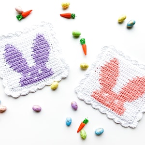 May include: Two crocheted bunny-shaped dishcloths, one purple and one pink, with white crochet edging. The dishcloths are surrounded by small plastic Easter eggs and carrots.