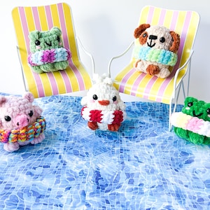May include: A collection of handmade crochet animals, including a frog, pig, dog, and chick, each wearing a colorful life preserver. The animals are arranged on a blue surface with a pool-like pattern, with two beach chairs in the background.
