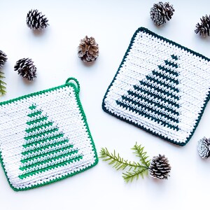 May include: Two green and white crocheted pot holders with a Christmas tree design. The pot holders are surrounded by pine cones on a white background.