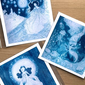 May include: Three square art prints featuring illustrations in shades of blue. One print depicts a woman in a flowing gown with a moon and stars. Another shows a mermaid-like figure in an underwater scene. The third print shows a woman in a flowing gown with a moon.