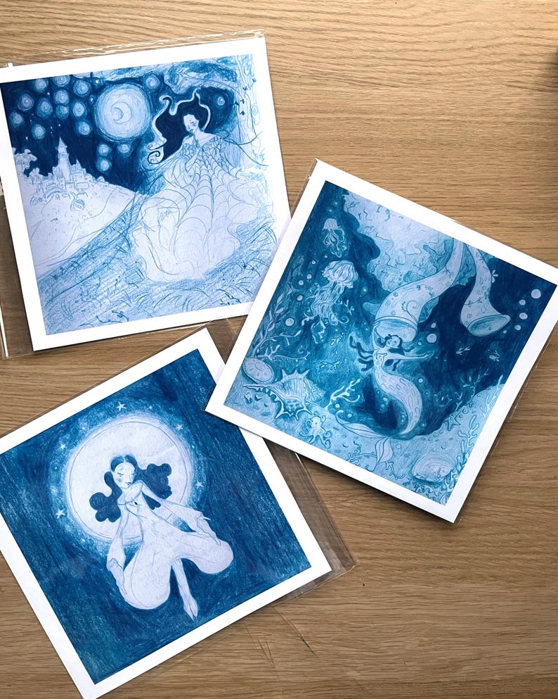 May include: Three square art prints featuring illustrations in shades of blue. One print depicts a woman in a flowing gown with a moon and stars. Another shows a mermaid-like figure in an underwater scene. The third print shows a woman in a flowing gown with a moon.