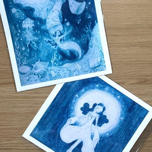 May include: Two blue-toned art prints featuring mermaid illustrations. One depicts a mermaid in an underwater scene with jellyfish and coral. The other shows a woman in a flowing gown in front of a moon with stars.
