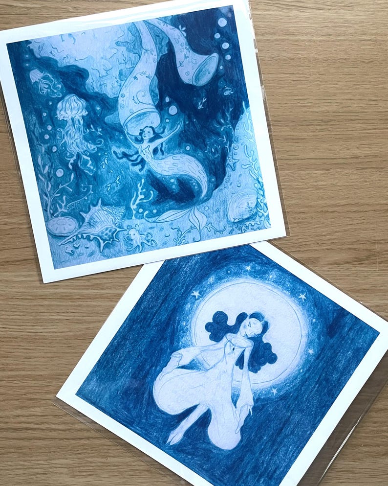 May include: Two blue-toned art prints featuring mermaid illustrations. One depicts a mermaid in an underwater scene with jellyfish and coral. The other shows a woman in a flowing gown in front of a moon with stars.