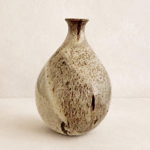 Könnte beinhalten: Eine Keramikvase mit bauchigem Körper und engem Hals. Die Oberfläche weist eine gesprenkelte Textur in Beige-, Creme- und Dunkelbrauntönen auf, was ein natürliches, erdiges Erscheinungsbild mit einer glatten, glänzenden Oberfläche erzeugt.