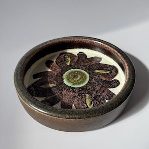 Peut inclure: Un bol rond en céramique avec un extérieur marron foncé et un motif floral. L'intérieur présente une base blanche avec un motif de fleur marron et un centre vert. Le bord est d'une nuance de marron plus foncée.