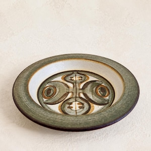 May include: A round ceramic plate featuring a wide, textured sage green rim and a central basin decorated with a symmetrical, abstract pattern in shades of olive green, brown, and cream. The plate has a low profile and a smooth, glazed finish.