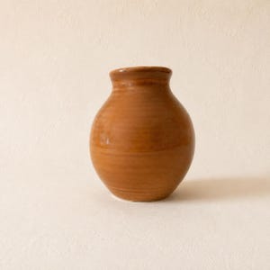 Könnte beinhalten: Eine kleine, runde Keramikvase mit einer glatten, glänzenden Oberfläche in einem warmen, erdigen Terrakotta-Braun. Das Gefäß hat einen kurzen, schmalen Hals, der sich am Rand leicht weitet, und einen bauchigen, kugelförmigen Körper.