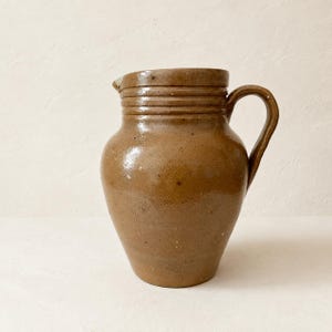 May include: A medium-sized, bulbous ceramic pitcher with a glossy, light brown glaze. The vessel features a wide, rounded body, a short neck with three horizontal incised rings, a small pouring spout, and a sturdy, curved handle on the right side.