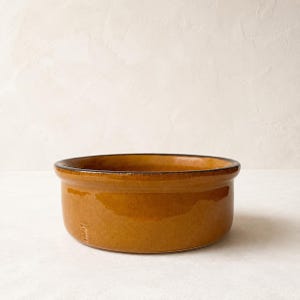 Może przedstawiać: Okrągła, niska miska ceramiczna z błyszczącym, ciepłym, ochrowo-żółtym szkliwem. Miska posiada gładki, lekko rozszerzający się brzeg oraz małą, wytłoczoną literę T na zewnętrznej ściance, co podkreśla jej prosty i funkcjonalny design.