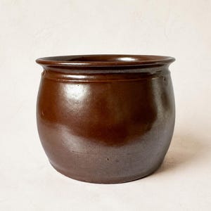 Może przedstawiać: Okrągły, ciemnobrązowy garnek ceramiczny o gładkim, błyszczącym wykończeniu i szerokim, lekko rozszerzającym się rancie. Naczynie ma pękaty korpus zwężający się ku podstawie, pokryty na całej zewnętrznej powierzchni jednolitą, ciemnobrązową glazurą.