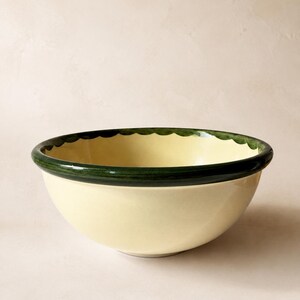 Puede incluir: Un cuenco redondo de cerámica con interior y exterior de color crema. El borde presenta una banda gruesa pintada de verde oscuro con un patrón decorativo festoneado en un tono verde más claro a lo largo de la circunferencia interior.