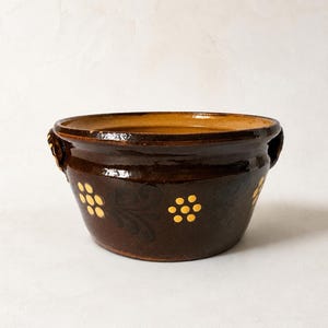 Può includere: Una ciotola in ceramica smaltata marrone scuro con due piccole maniglie laterali e un motivo decorativo di fiori dipinti in giallo, ciascuno con sei petali. L'interno della ciotola presenta uno smalto color beige più chiaro e lucido.