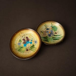 Può includere: Due piattini rotondi in ceramica con una smaltatura di base gialla. Ogni piattino presenta disegni astratti dipinti a mano in blu, verde, marrone e nero, tra cui forme a stella, motivi a griglia e pennellate. I bordi sono marrone scuro e rialzati.