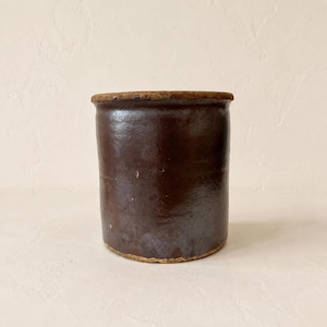 May include: A cylindrical stoneware crock featuring a deep, dark brown glossy glaze. The vessel has a thick, slightly textured rim and a flat base, with subtle variations in the glaze finish across the exterior surface of the ceramic container.