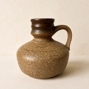 Könnte beinhalten: Ein Keramikkrug mit bauchigem Körper und engem Hals. Das Stück zeigt eine gesprenkelte braune Glasur im unteren Bereich, die in ein glattes, dunkelbraunes Finish am oberen Hals und Rand übergeht. Es verfügt über einen einzelnen geschwungenen Henkel.