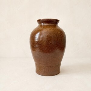 Könnte beinhalten: Eine mittelgroße Keramikvase mit bauchigem Körper und engem Hals. Die Außenseite ist mit einer glänzenden, gesprenkelten braunen Glasur überzogen, die durch subtile Farbvariationen eine texturierte Optik auf der gesamten Oberfläche erzeugt.