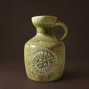Pode incluir: Um jarro de cerâmica com um esmalte verde claro texturizado e salpicado. O corpo apresenta um padrão floral circular em relevo na parte frontal. Possui um gargalo curto e largo, uma borda alargada e uma asa curva fixada na lateral.