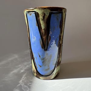 Può includere: Un vaso in ceramica fatto a mano con un design unico. Il vaso presenta un interno blu con un contorno marrone e un esterno verde e marrone. Il vaso ha una forma cilindrica alta e viene probabilmente utilizzato per scopi decorativi.