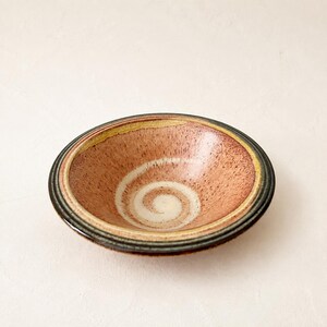 Könnte beinhalten: Eine flache Keramikschale mit gesprenkeltem terrakotta- und cremefarbenem Innenraum und einem markanten weißen Spiralmuster in der Mitte. Der Rand ist durch konzentrische Bänder in Gelb, Dunkelgrün und Schwarz definiert, die einen Schichteffekt erzeugen.