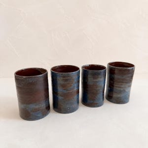 Puede incluir: Cuatro vasos cilíndricos de cerámica dispuestos en fila. Cada recipiente presenta un esmalte moteado aplicado a mano con tonos mezclados de marrón oscuro, azul pizarra y gris carbón, creando una apariencia texturizada y en capas.