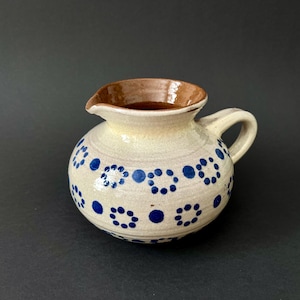 Può includere: Piccolo brocca rotonda in ceramica con manico e beccuccio. La brocca è bianco sporco con bordo e manico marroni, e decorata con motivi a pois blu. Probabilmente utilizzata per servire liquidi.