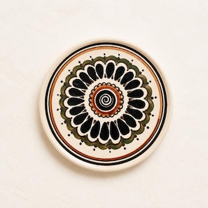 Könnte beinhalten: Ein runder Keramikt runder Keramikt runder Keramikteller mit einer zentralen schwarzen Spirale, umgeben von konzentrischen Ringen aus orangefarbenen, grünen und schwarzen blütenblattartigen Formen. Das Design ist symmetrisch, mit cremefarbenem Hintergrund und dünnen schwarzen Kreislinien.