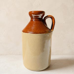 Può includere: Una brocca in ceramica con un design a due tonalità. La parte superiore, compresi il collo e il manico, è rifinita in marrone scuro lucido, mentre il corpo cilindrico presenta una smaltatura beige chiaro liscia con una superficie riflettente.